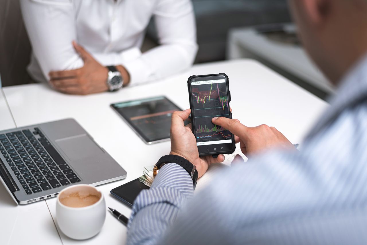 Two businessmen examine stock market data on a smartphone and laptop in a modern office setting.
